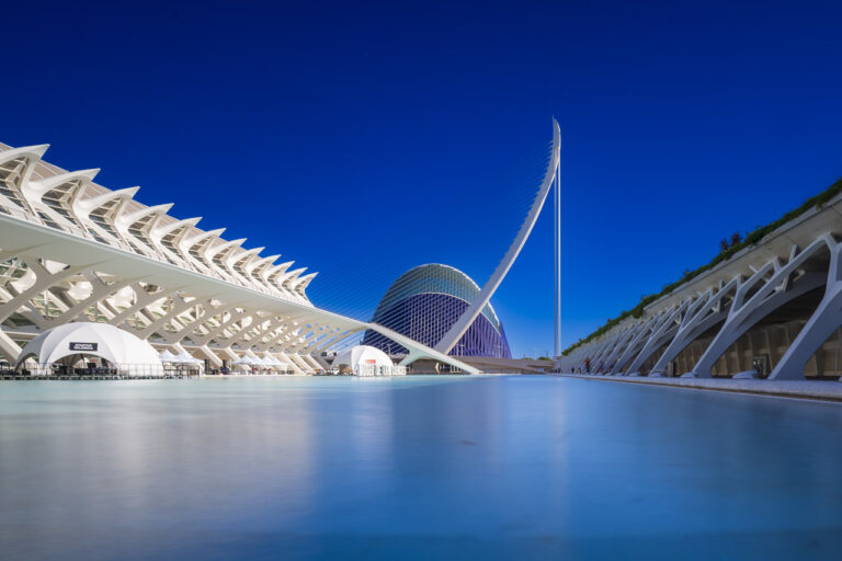 The Museu de les Ciències Príncipe Felipe, and the water surrounding it, with the Pont l’Assut de l’Or bridge and CaixaForum in the background and clear blue skies.