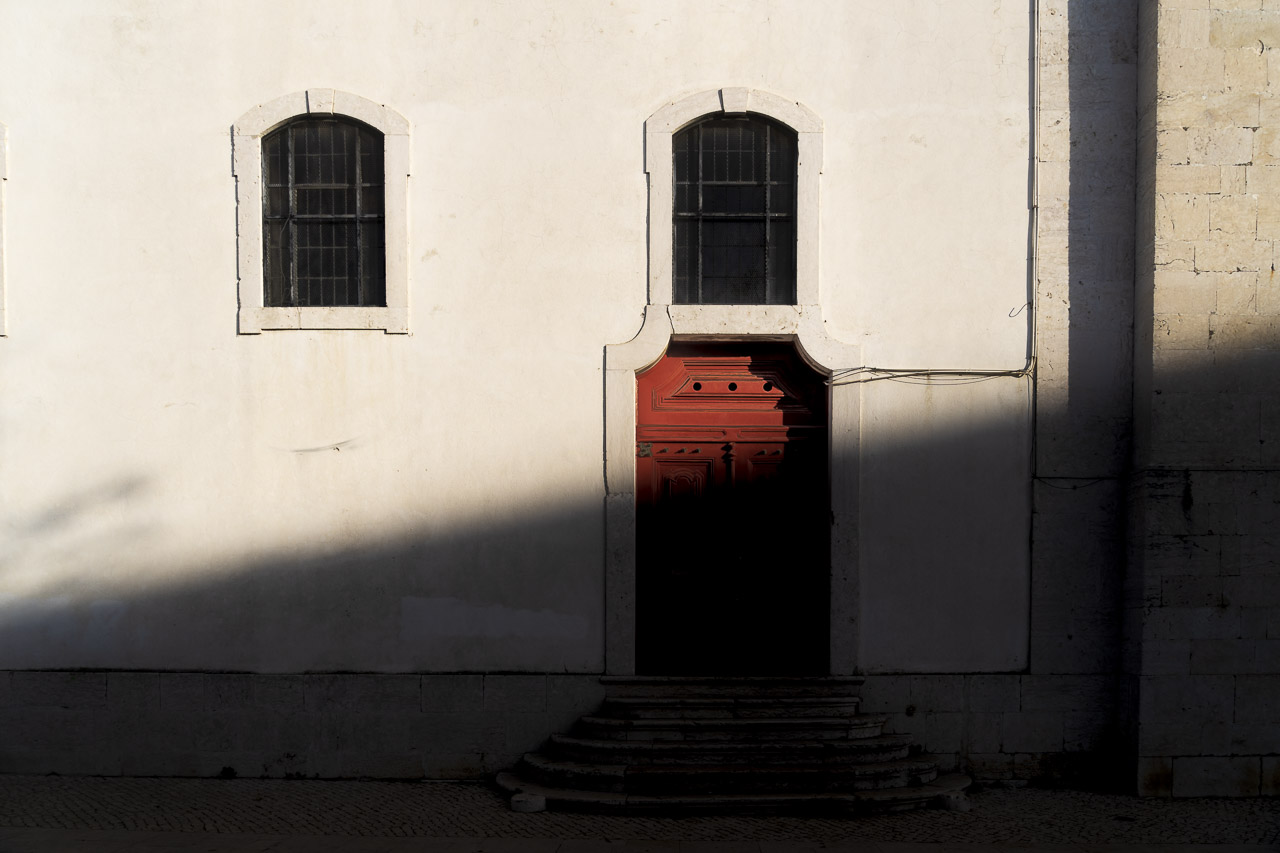 A side door of the Igreja Paroquial da Graça in Lisbon catches the first rays of the sun. The door is red, and a long diagonal shadow stretches across the door.