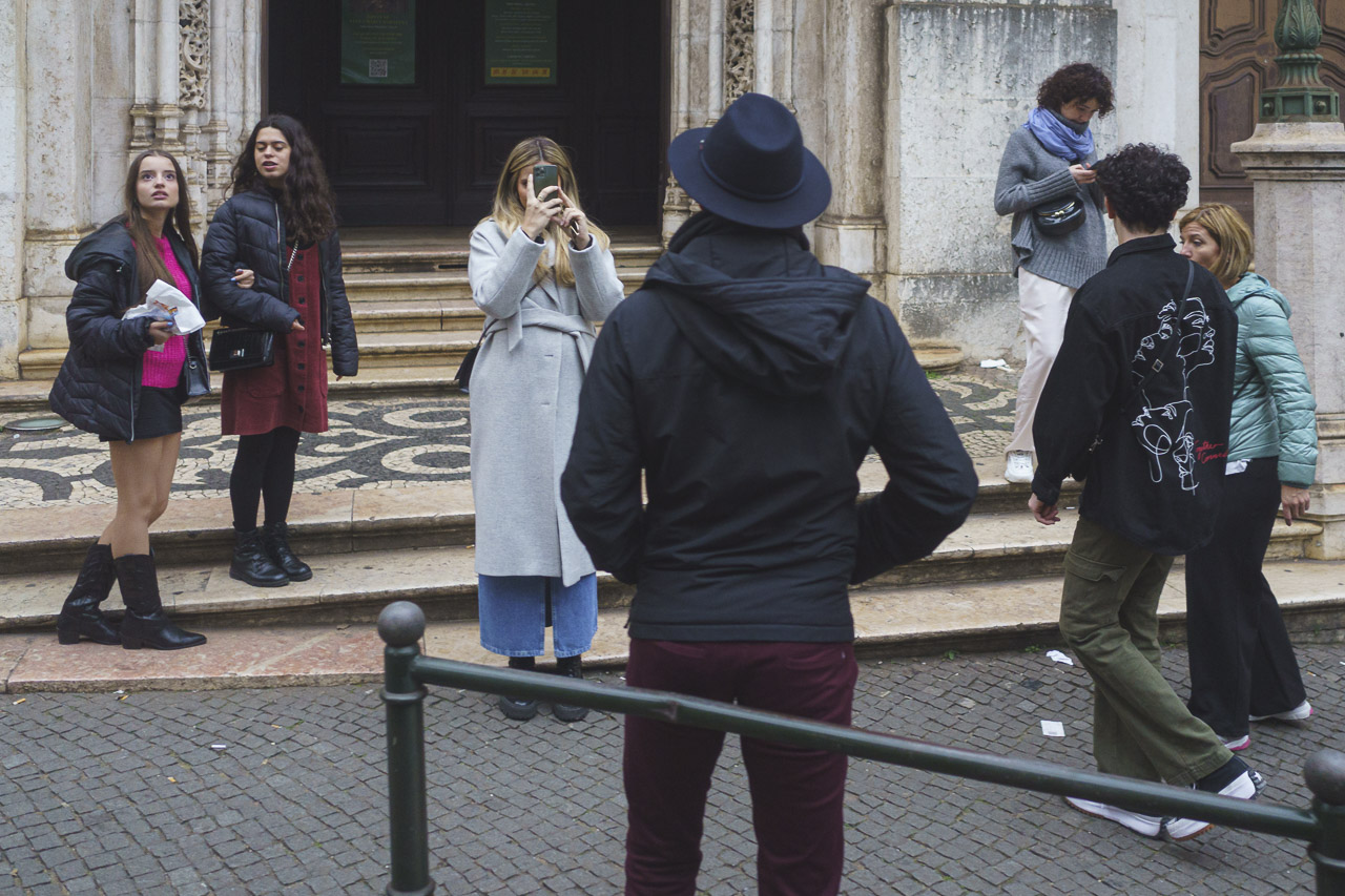 A woman photographs a man in a hat, in front of the tram I'm on. She has her phone up to her face. 2 younger women are nearby, while a couple walk in from the right and a woman looks at her phone in the background near them.