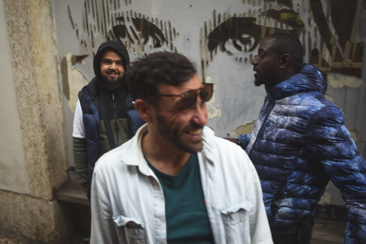 Three men pictured on a street. Two of them smiling while the third is walking past. In the background is a large torn poster with eyes visible.