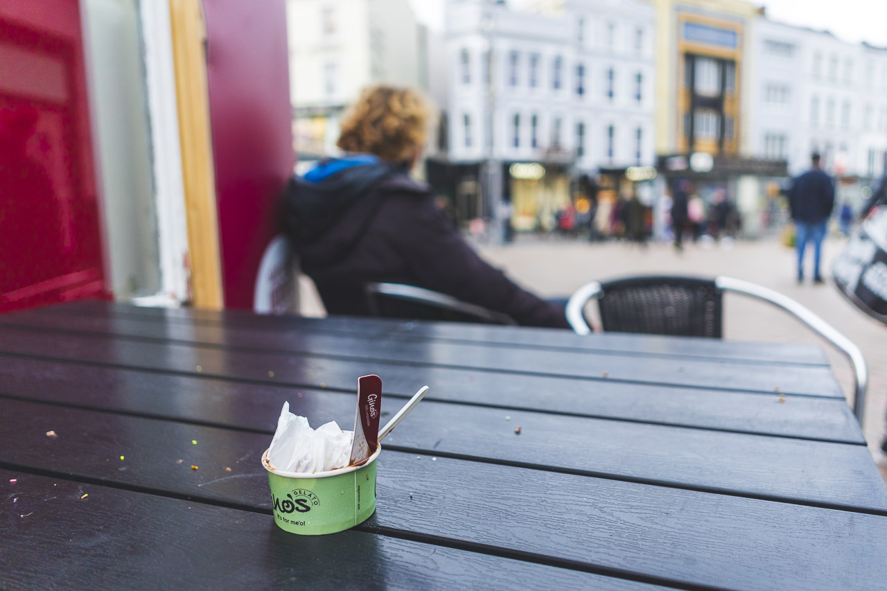 An outside table in a city with a man sitting, facing the other way. A used tub of ice cream sits on the table with paper, wrapper and a fork in it.