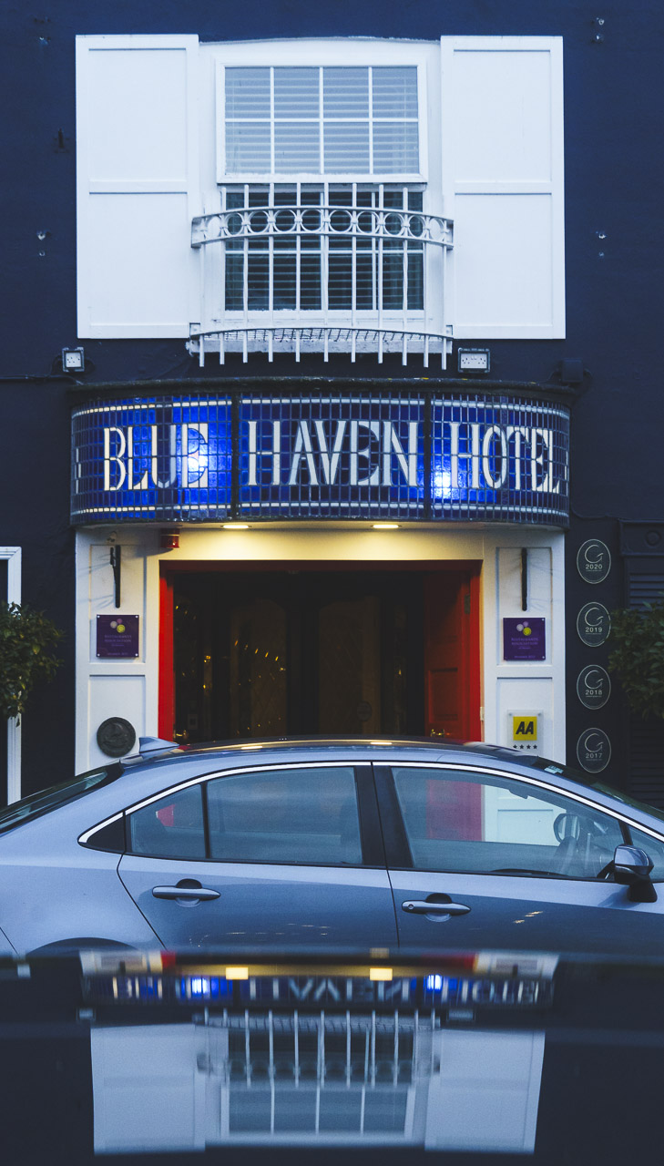 The entrance to the Blue Haven Hotel, reflected in a nearby parked car. The window above has a white frame, while the name of the hotel sits above the main door in a beautiful blue and white glass piece.