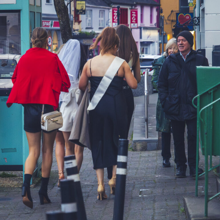 Four women dressed to go out on a hen night as one of them is wearing a wedding veil and another is wearing a sash. They are watched by an older couple as they walk by on the street.