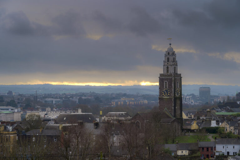 The spire of St. Anne's Church rises above nearby homes. The Firkin Crane is seen nearby, and the County Hall is far off in the distance. A crack in the cloud cover at the horizon allows in a smattering of light from the setting sun.