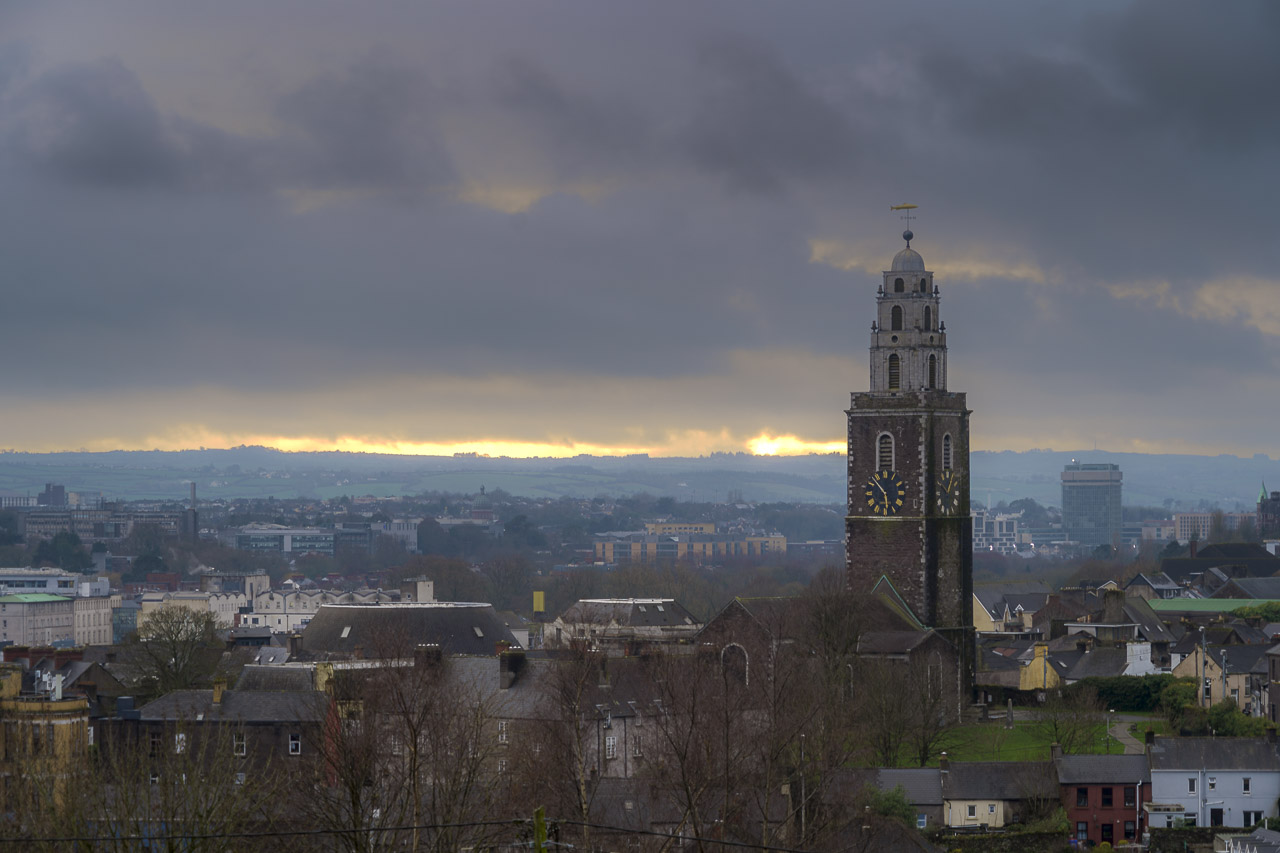 The spire of St. Anne's Church rises above nearby homes. The Firkin Crane is seen nearby, and the County Hall is far off in the distance. A crack in the cloud cover at the horizon allows in a smattering of light from the setting sun.