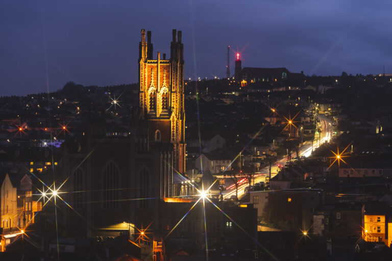 The Cathedral of St Mary & St Anne, or North Cathedral, in Cork sits on Cathedral Road where the light streaks of passing traffic can be seen. The Church of the Ascension is seen at the top of the photo, with its distinctive red cross glowing in the dark. Point lights make star shapes in the photo.