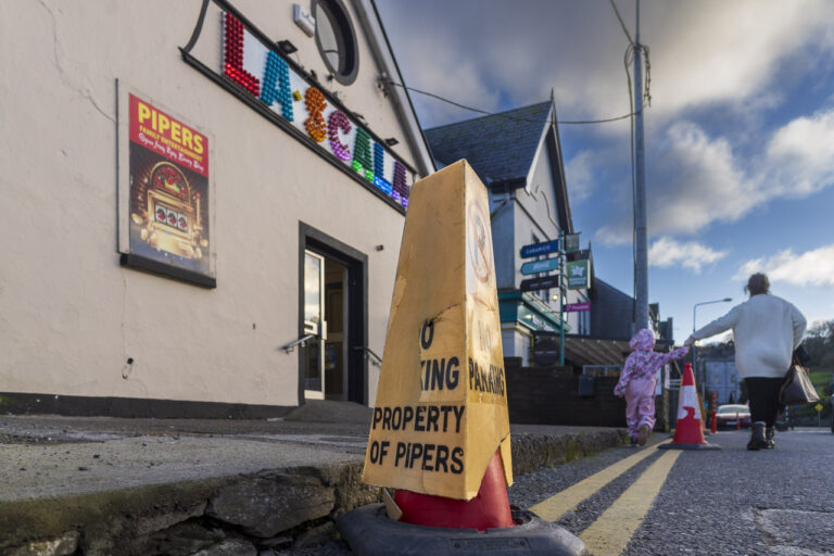 A "no parking" bollard placed at the side of the road in Crosshaven proclaims it is the property of Pipers. That's a fairground in the town. A mother and daughter hold hands and walk into the background. La Scala is seen in the image too.