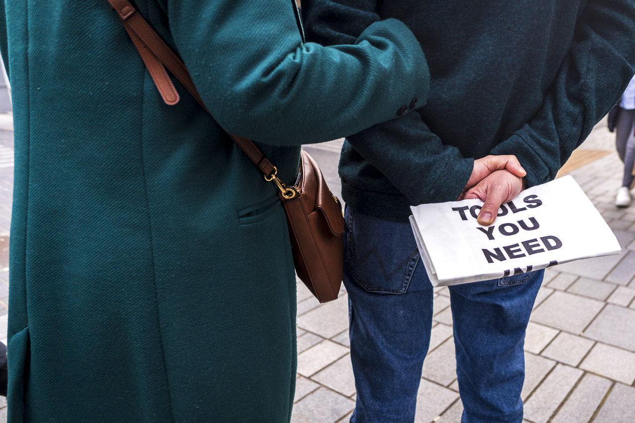 A man holds a paper bag with the text, "TOOLS YOU NEED" on it, while a woman holds his arm. She is carrying a handbag. Picture from behind both.