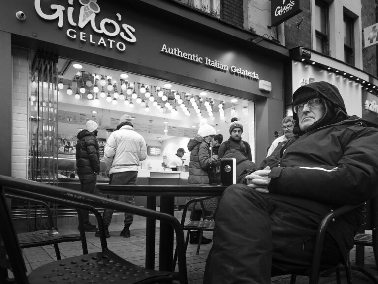 A man dressed warmly in a coat, with a hood over his head sits at a table outside Gino's Gelato. He's listening to a radio while other people