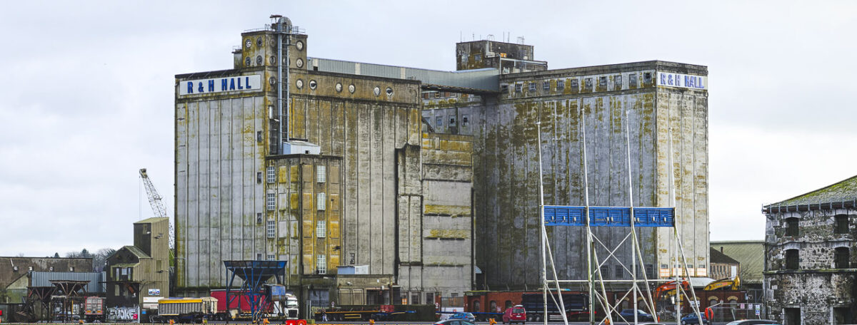 The large R&H Hall building on Kennedy Quay in Cork. The back of the Welcome to Cork" sign is visible in the foreground. Other buildings and vehicles can be seen too, tiny in comparison.