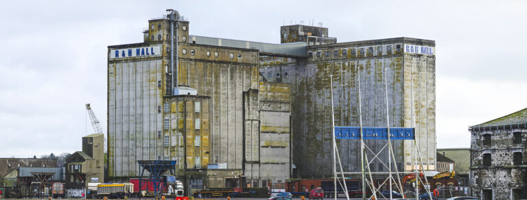 The large R&H Hall building on Kennedy Quay in Cork. The back of the Welcome to Cork" sign is visible in the foreground. Other buildings and vehicles can be seen too, tiny in comparison.