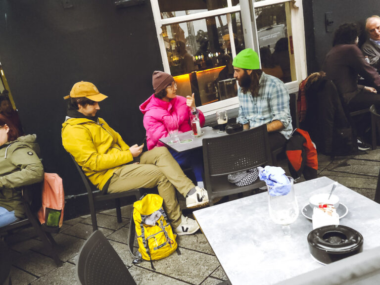 3 people sitting at a table outside a pub wearing bright clothes. A man wearing yellow. A woman wearing a bright pink jacket. Another man wearing a bright green woolly hat. People around them wearing ordinary, by contrast, drab clothing. Another table sits empty in front of them.