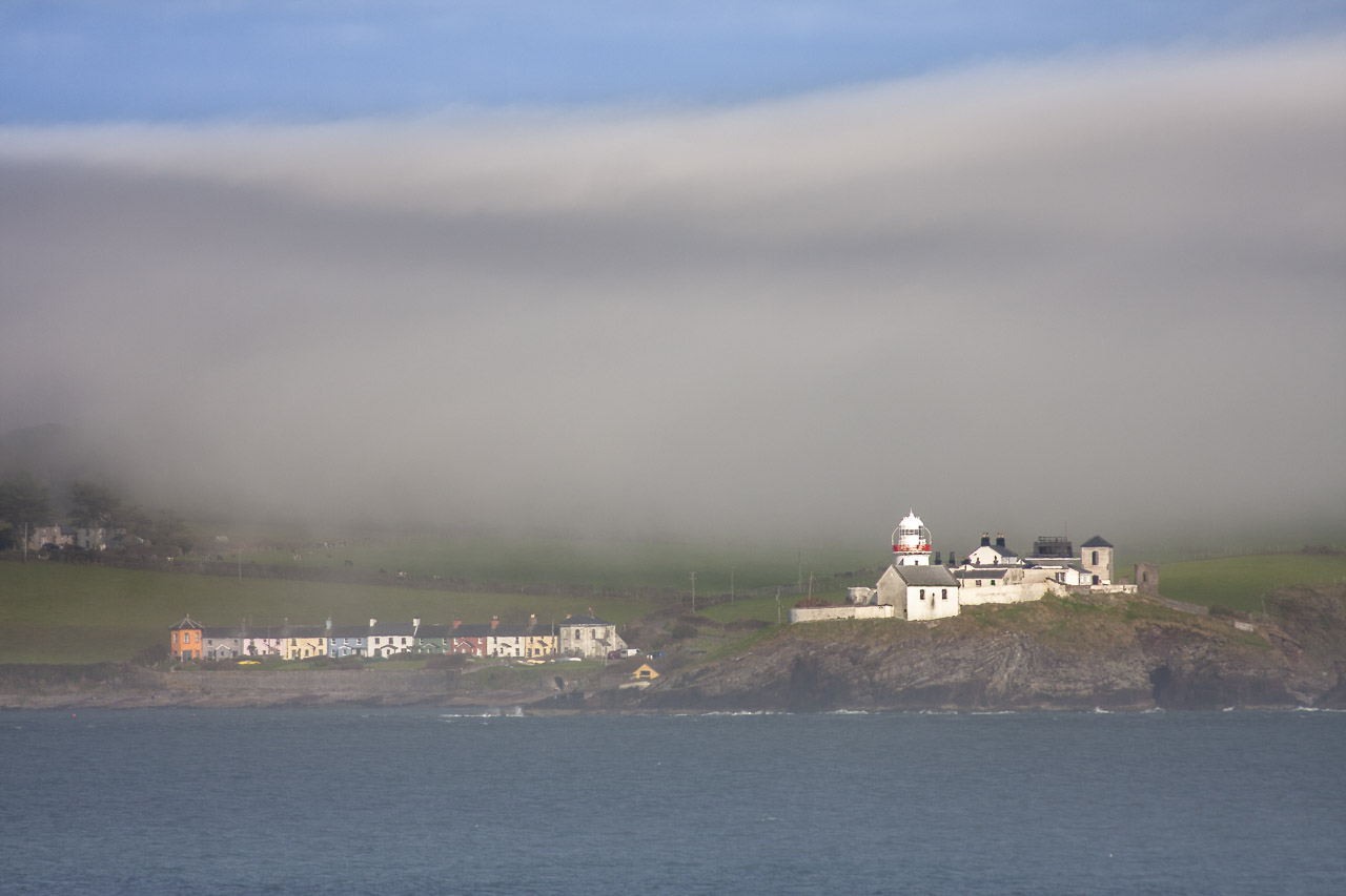 Fog flows over the landscape and will soon cover the small town and the lighthouse. The water of Cork Harbour can be seen in the foreground.