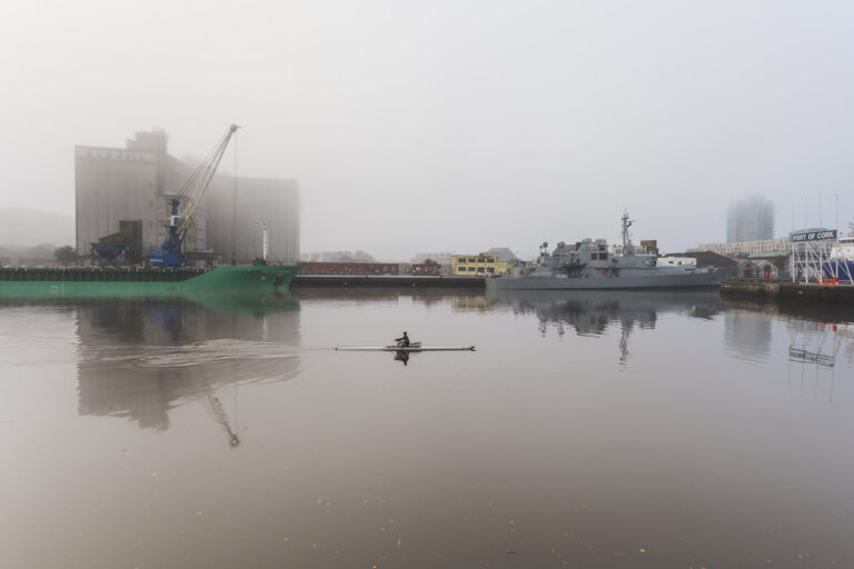 A man in a solitary boat rows up a river past ships docked at the Quay. Buildings in the background are somewhat obscured by fog, and the "Port of Cork" sign is visible to the far right of the image.