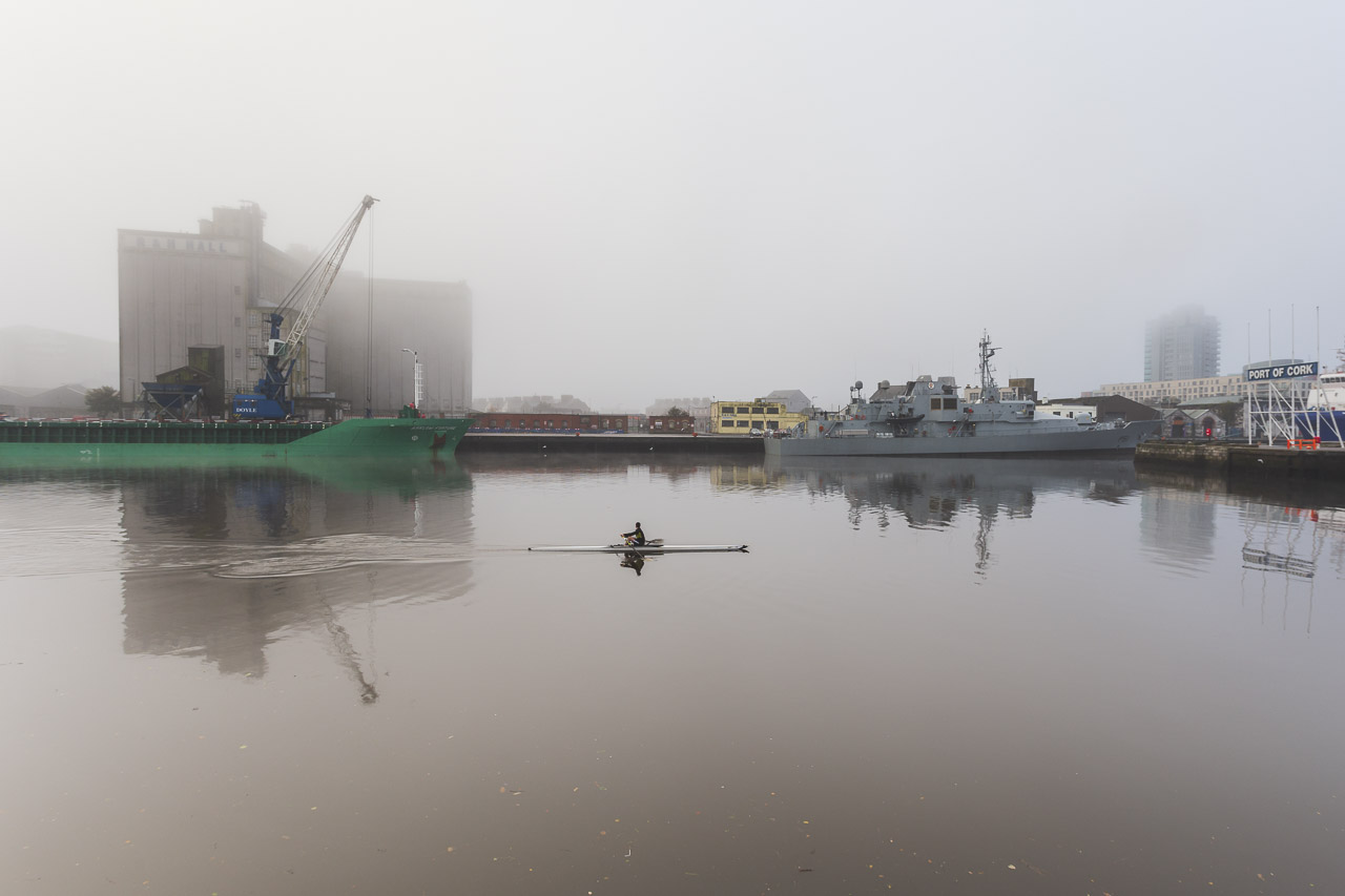 A man in a solitary boat rows up a river past ships docked at the Quay. Buildings in the background are somewhat obscured by fog, and the "Port of Cork" sign is visible to the far right of the image.