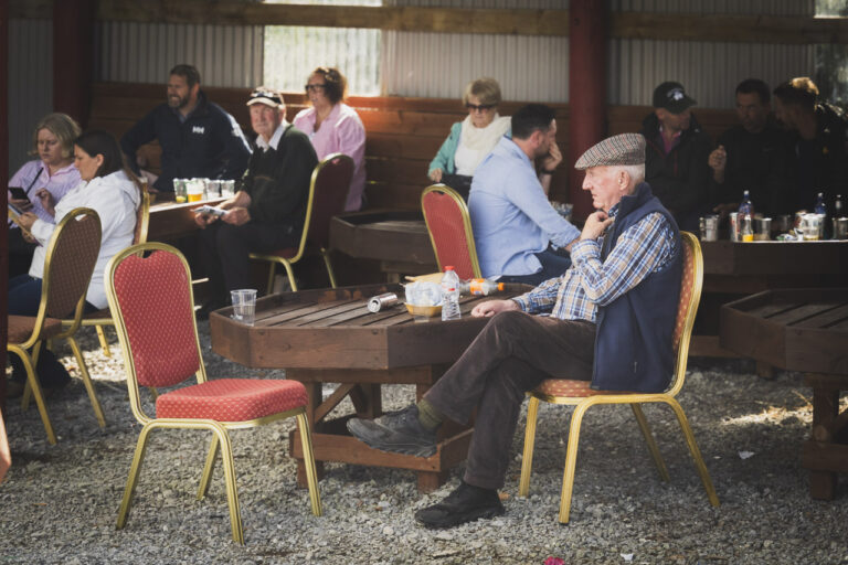 A man sits at a table surrounded by other tables with people at them. He is alone, facing an empty chair.