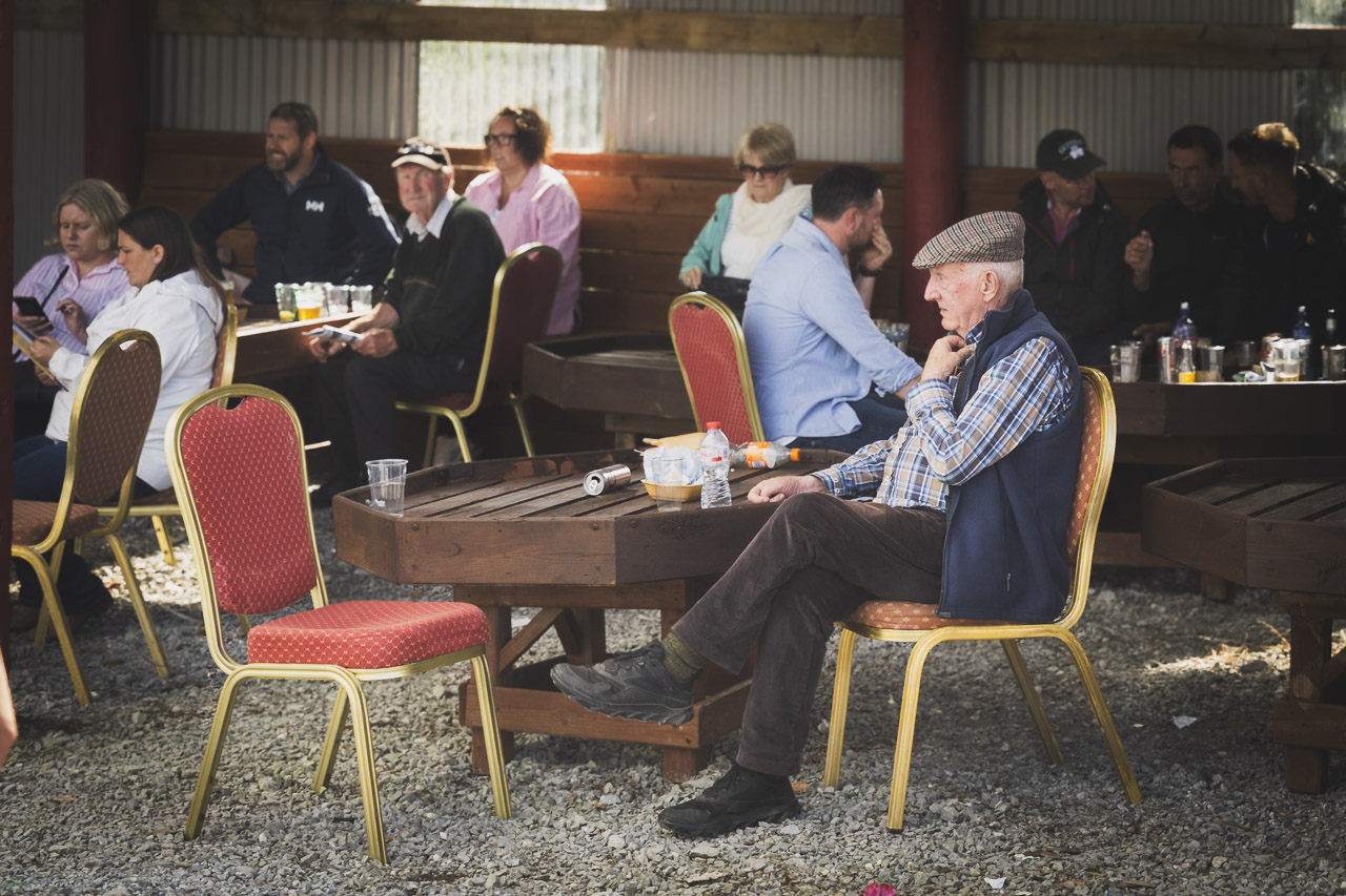 A man sits at a table surrounded by other tables with people at them. He is alone, facing an empty chair.