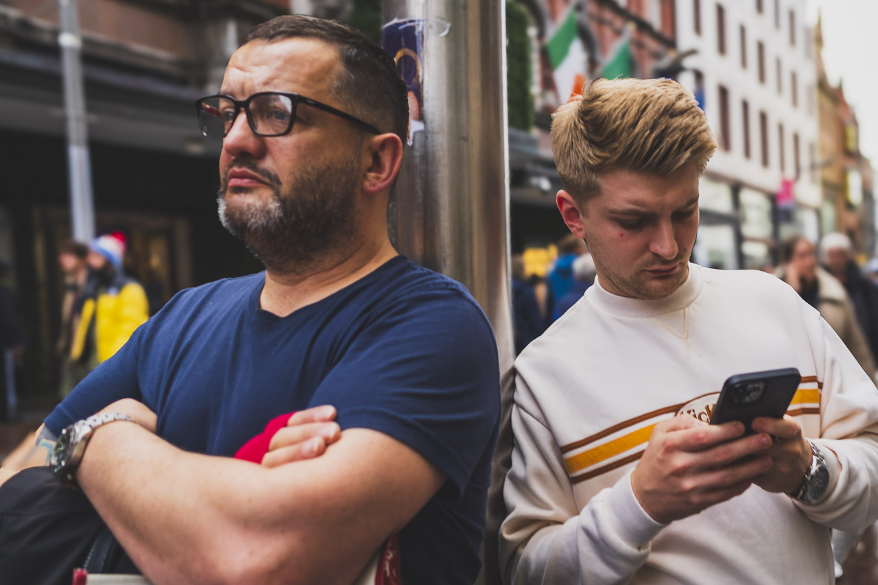 Two men lean against a pole. One checks his phone, while the other one looks off in the distance.