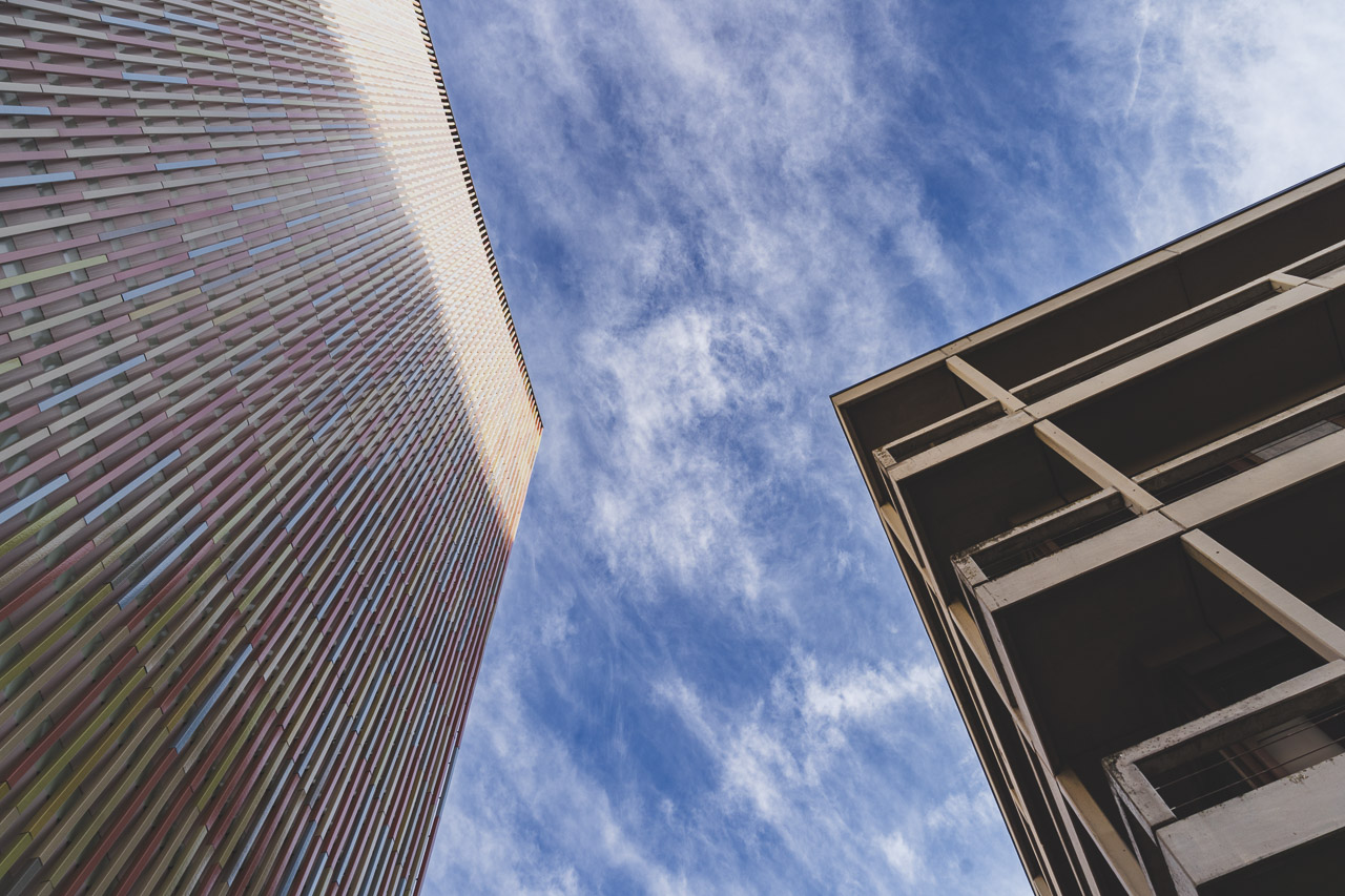 Looking up next to a building with coloured stripes to the left, and an apartment block to the right. Blue sky with light cloud in the middle.