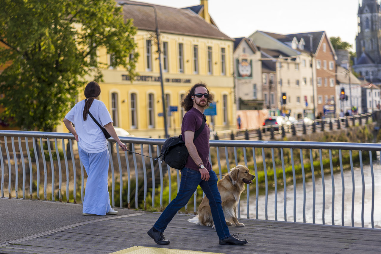 A woman holding her dog on the lead. He's sitting on the ground of a pedestrian bridge. The river visible in the background. A man walks past, looking directly at me.