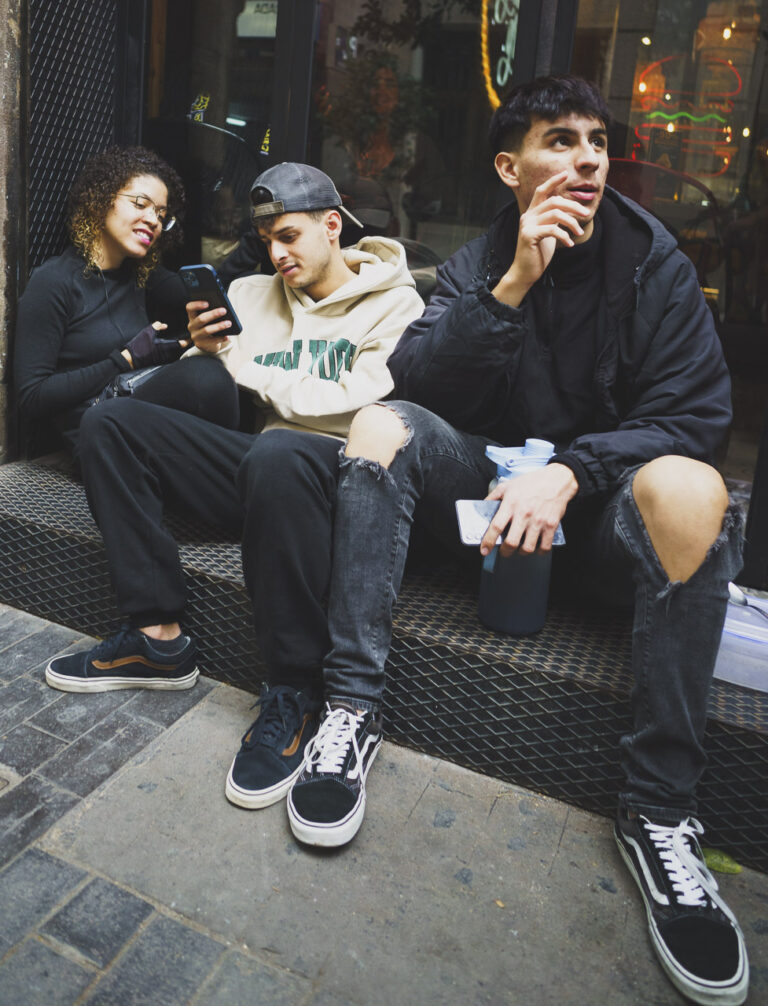 A group of young people sitting on a window sill down a side street. A woman and two men. They each have phones and the man on the right is speaking to someone outside the frame.