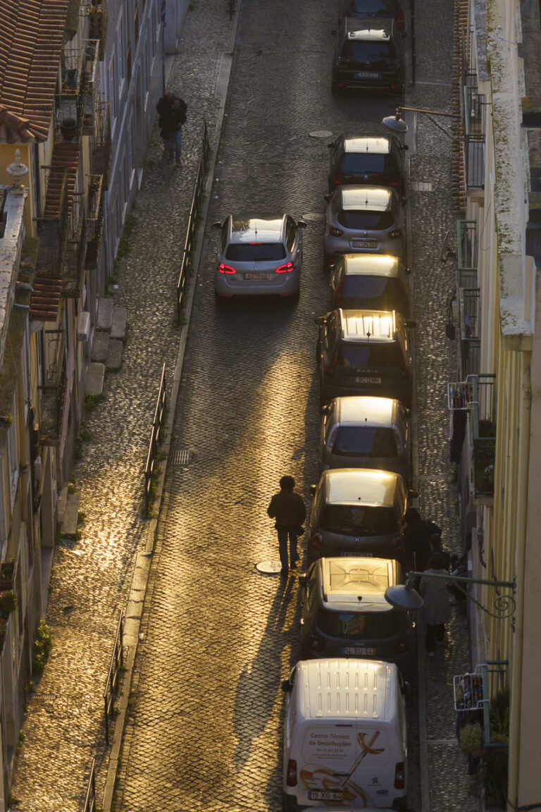 A street at sunset from above. A person walks in the light of the setting sun, casting a long shadow behind them. Another figure walks up the road, while a group of people are on a path to the right. Lots of parked cars and the brake lights of a car on the right are visible as it is on a hill.