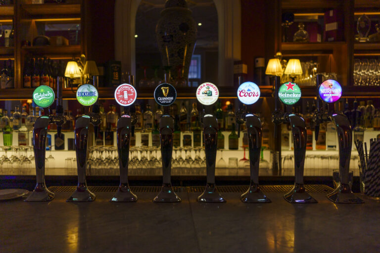 At the bar of a pub in a hotel. The taps that pour the drinks are labelled with the brands of the drinks offered. Carslberg, Rockshore, Smithwicks, Guinness, Birramoretti, Coors, Heineken, Murphys.