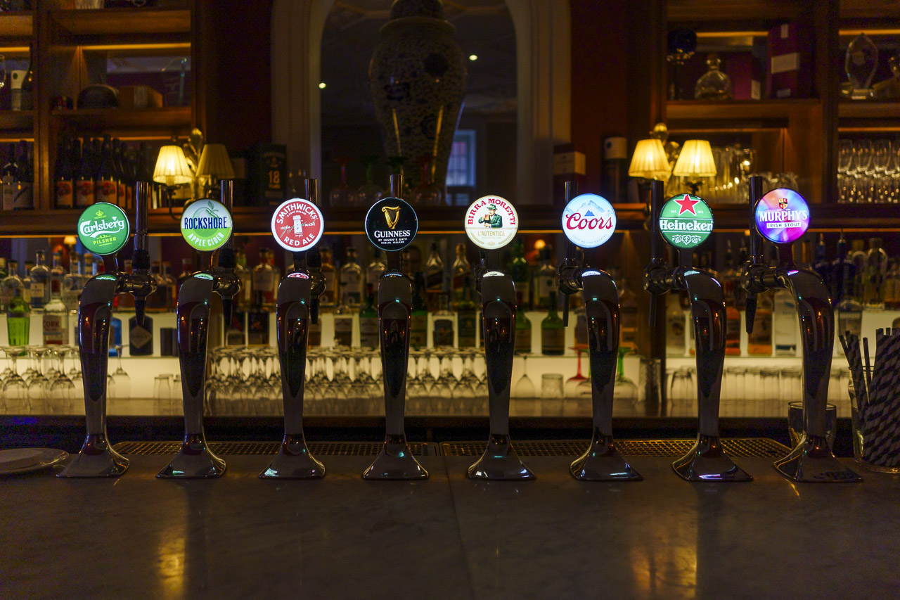 At the bar of a pub in a hotel. The taps that pour the drinks are labelled with the brands of the drinks offered. Carslberg, Rockshore, Smithwicks, Guinness, Birramoretti, Coors, Heineken, Murphys.