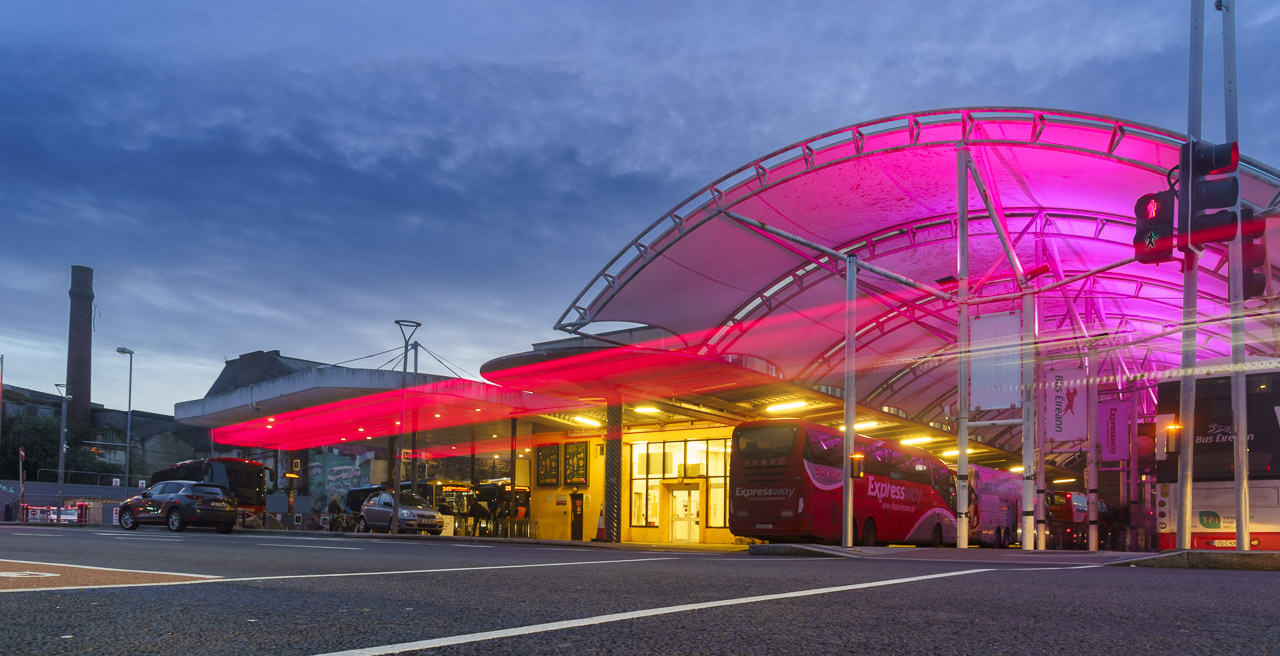 The roof of the bus station in Cork City is lit up by pink lighting as a vehicle drives past and brake lights are captured in mid air. Behind the bus station is the red brick chimney stack belonging to a factory that sat in that location from around 1920 but was demolished in recent years.