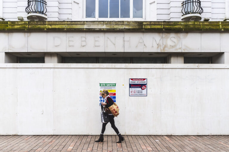 A woman walks past the front entrance of the old Roches Stores, or later Debenhams, but now a derelict building in the heart of the city. The building is surrounded by hoarding.