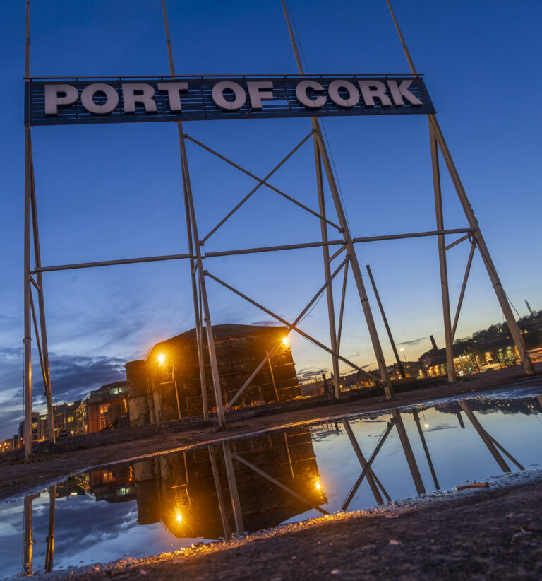 The sign saying "PORT OF CORK" displayed on the middle island of the city, visible to any boats coming up the river from the harbour. The supports of the sign are reflected in a puddle, as are the lights and nearby building. The Cork skyline is visible in the background of this sunset photo.