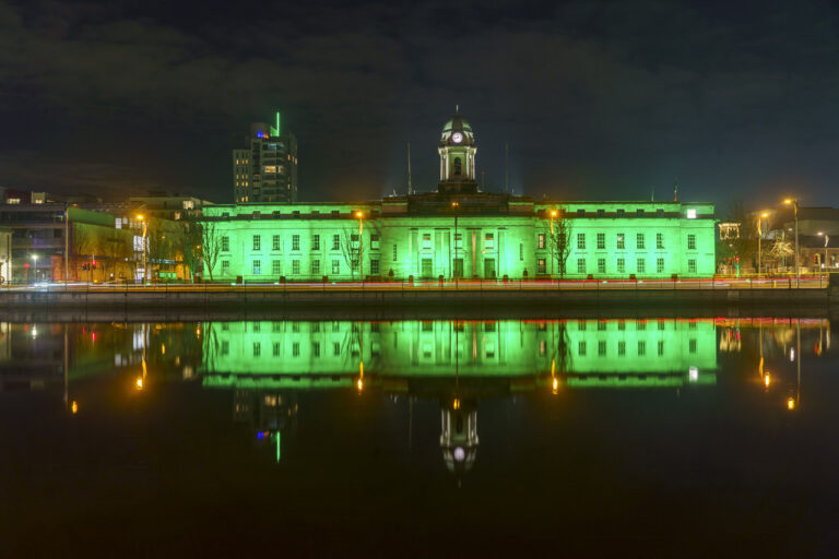 Cork City Hall at night, and it's lit up in green to celebrate St. Patrick's Day. The river reflects the colour of the building.