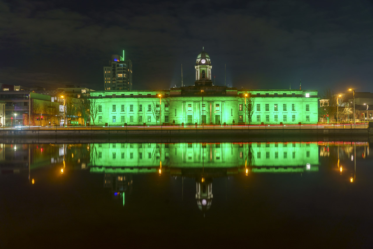 Cork City Hall at night, and it's lit up in green to celebrate St. Patrick's Day. The river reflects the colour of the building.