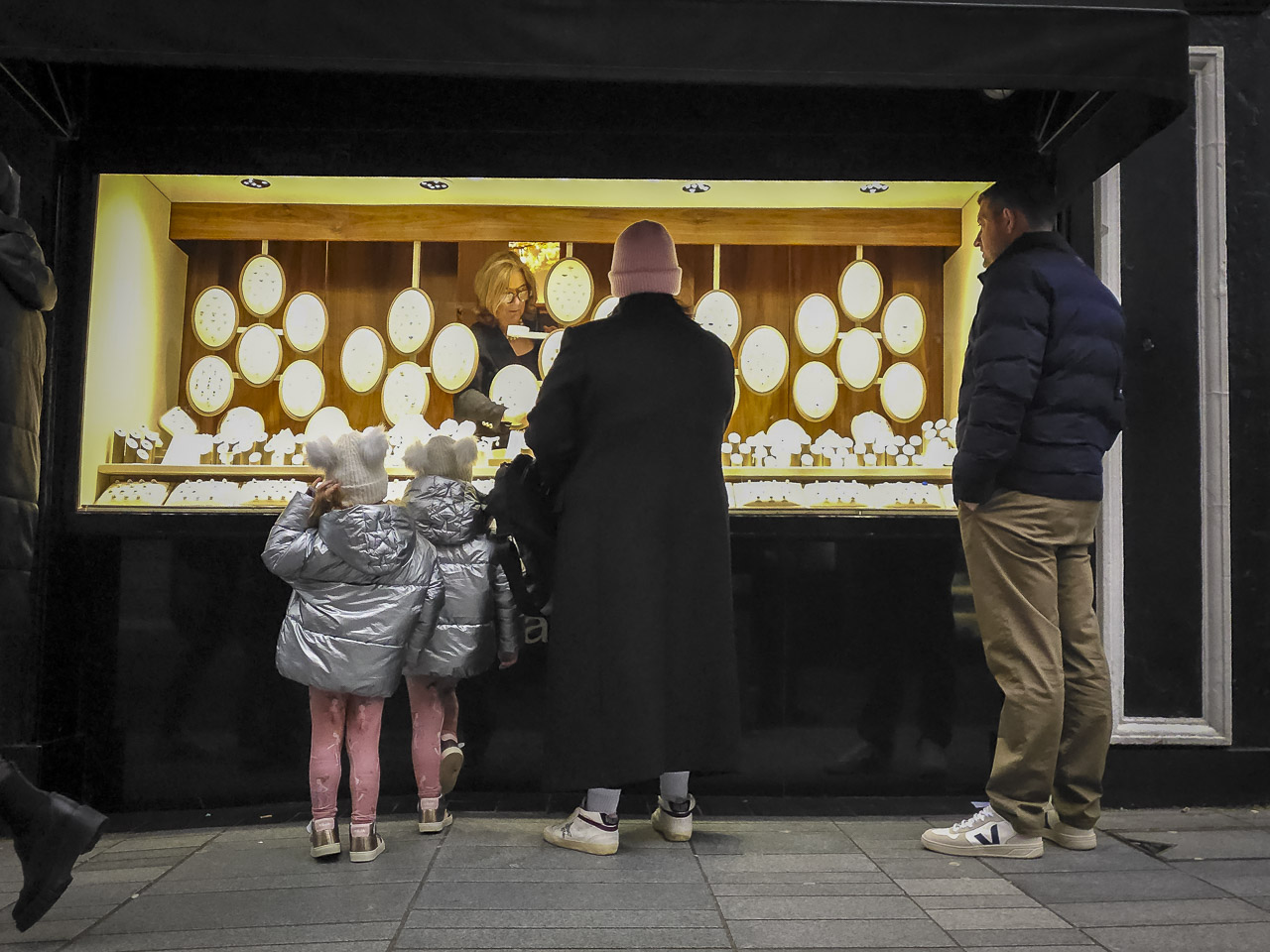 A woman and her two kids, with a man nearby, presumably her partner, look in the window of a jeweller's as someone inside picks out something.