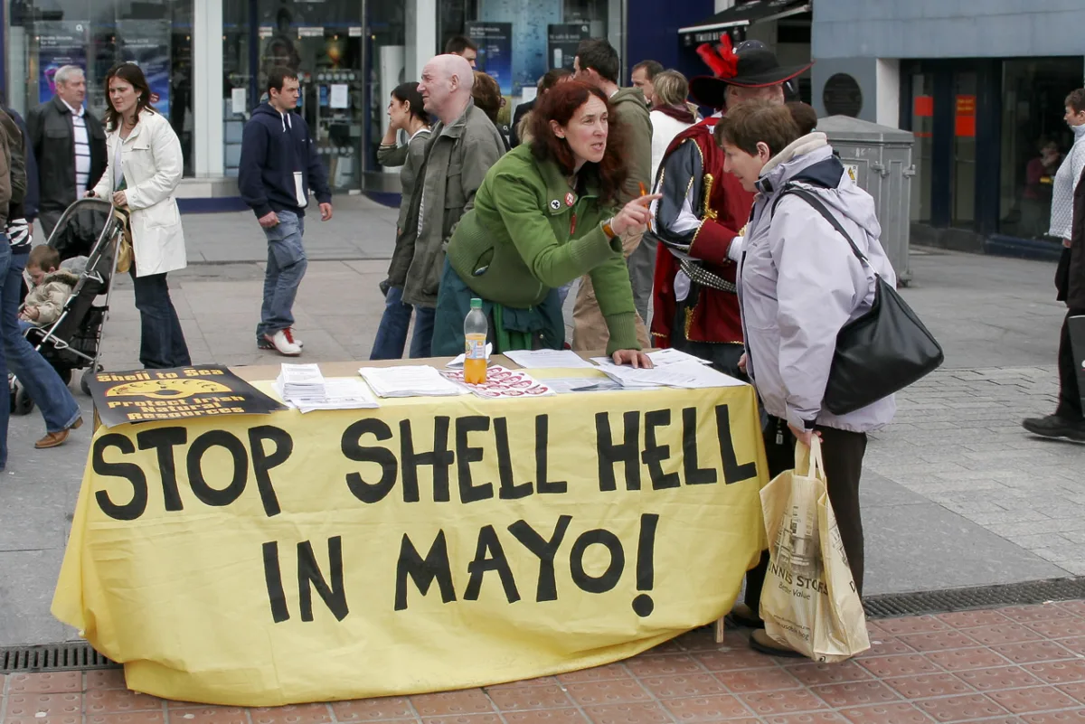 A woman stands behind a desk with the text, "Stop Shell Hell in Mayo!" printed on a covering on it. She is talking to a passer by, attempting to get a signature. Other people walk behind.