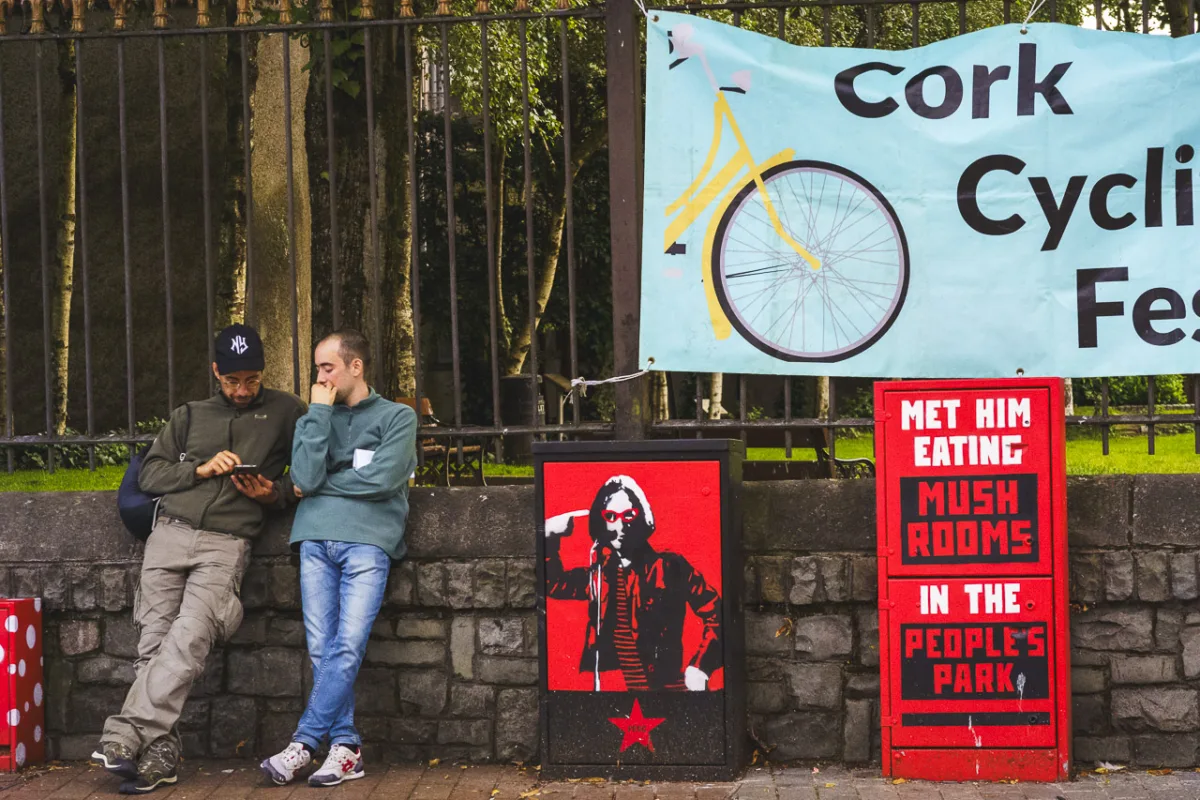 Two men lean against a wall while one checks his phone and the other looks on. A poster on railings behind them is partially seen and says "Cork Cycli.. Fes..." Street furniture is painted with the text "MET HIM EATING MUSH ROOMS IN THE PEOPLE'S PARK" and the other has a figure pointing at their own head with a mic on a stand.