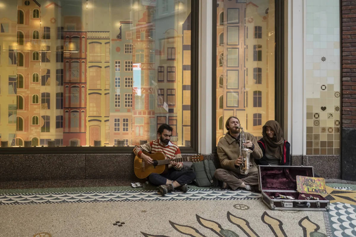 Three people sitting against a wall. Two are playing instruments, the guitar and saxaphone and the other person is smiling and enjoying the music. They have a box in front of them with a sign saying, "YOU ARE LOVED".