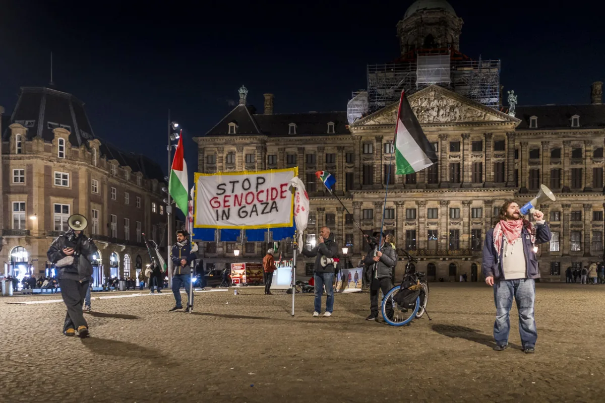 A protest in Dam Square, Amsterdam, against the genocide in Gaza by Israel. Protestors hold a sign saying, "STOP GENOCIDE IN GAZA" and two others shout into megaphones.