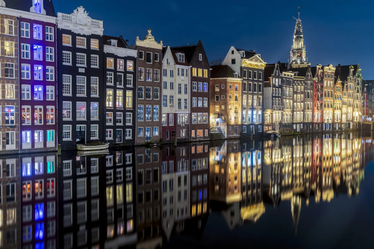 Houses by the water's edge are lit up and reflect in the still water. It's a night shot.