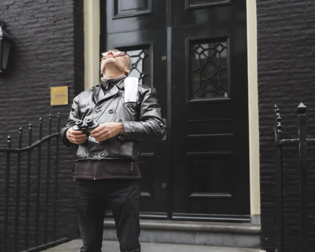 A man holding binoculars looks up at a building opposite him on a street.