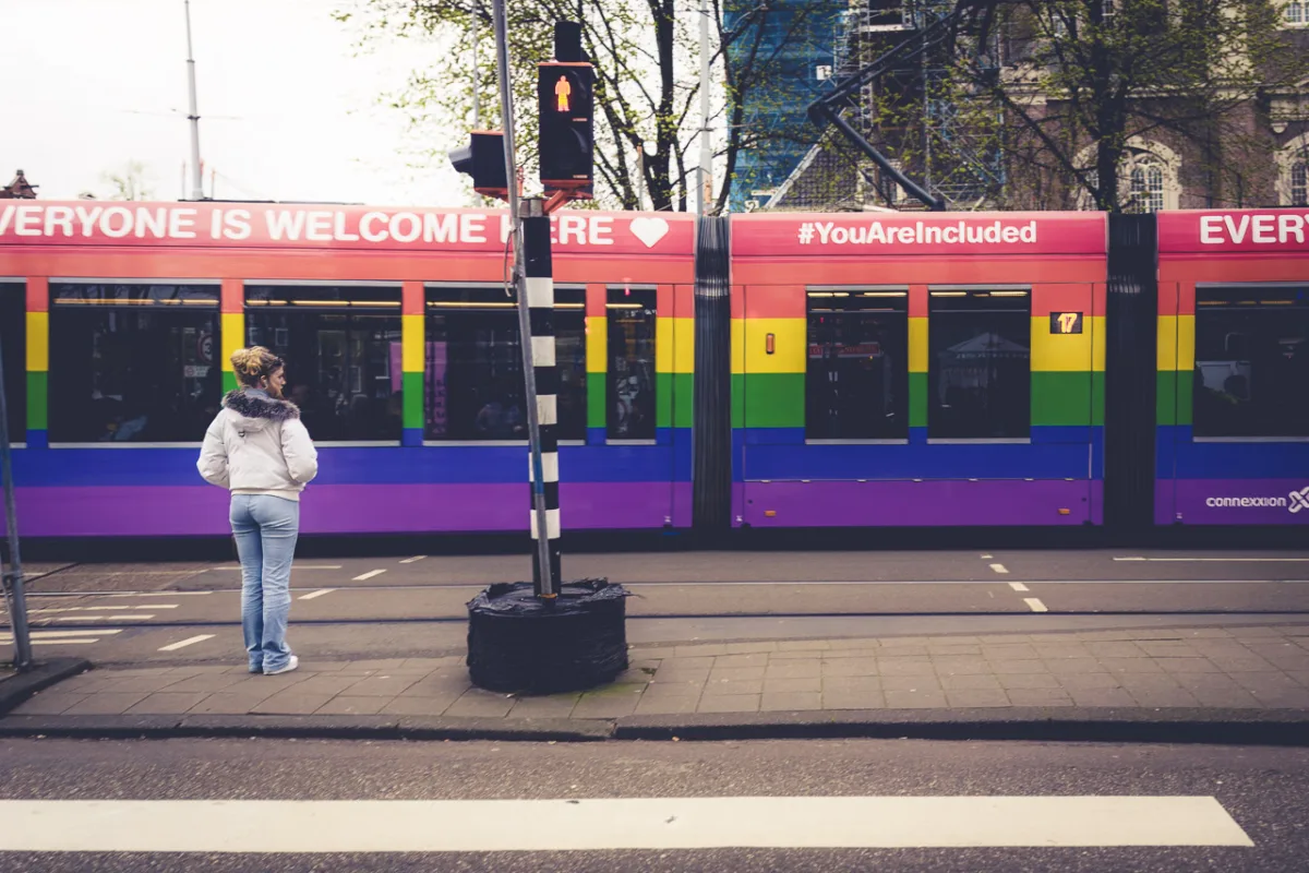 A train painted in the colours of the Pride Flag with the text "EVERYONE IS WELCOME HERE #YouAreIncluded" printed on it. A young woman waits to cross the road.