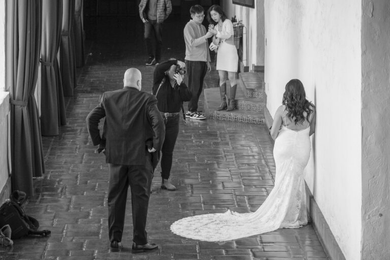 A photographer takes a photo of a bride in a corridor while her new husband looks on, and some tourists in the background film with a smartphone. A man is walking into the frame from the back.