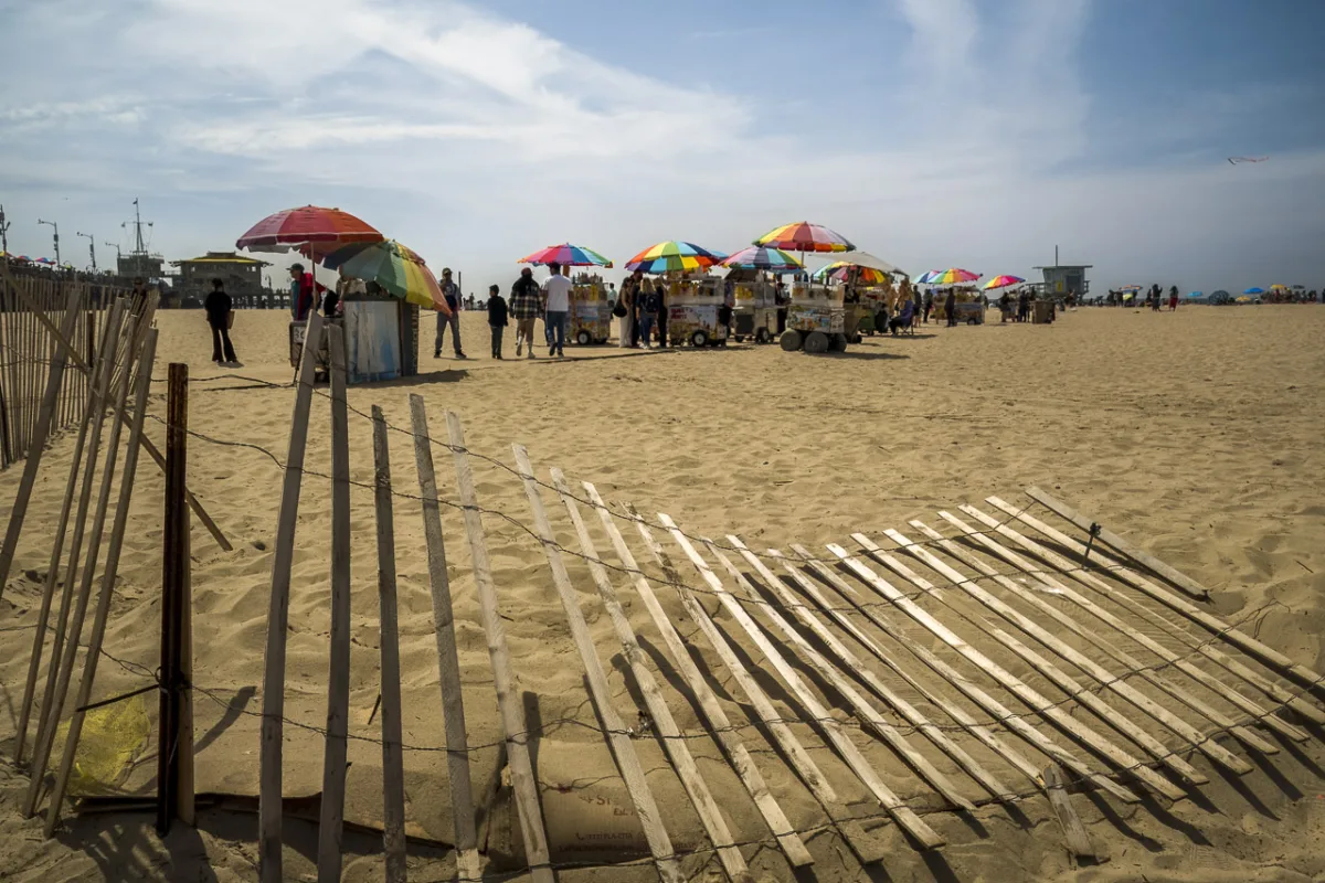 On a beach, a wooden fence lies partly on the ground. Halfway through the photo are lots of food carts surrounded by people. A pier is visible on the left and a lifeguard tower on the right, under a blue sky lined with clouds.