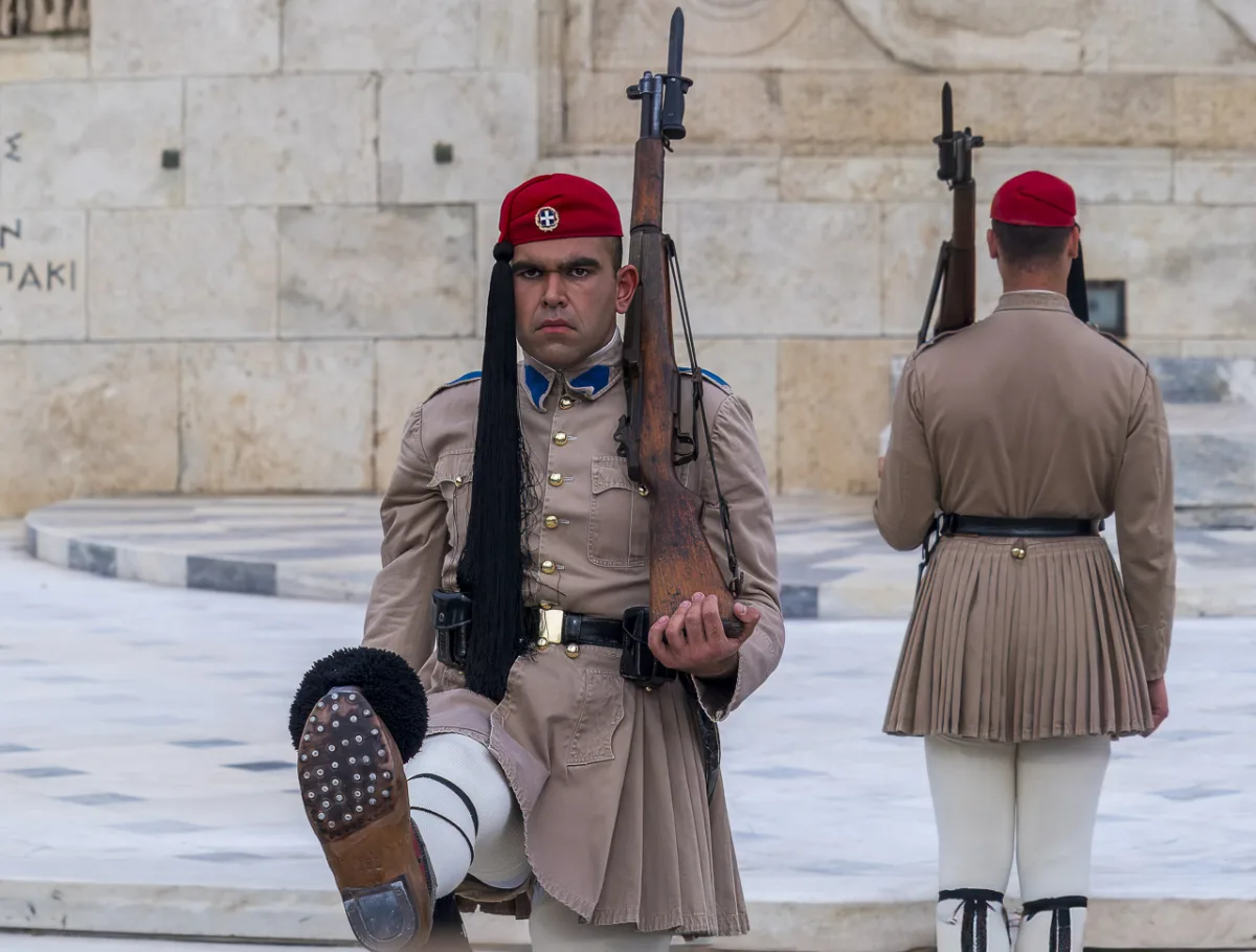 The changing of the guard. A guard approaches the camera. His dress uniform is a red cap with a long black silk tuft. His uniform is brown and his shoes have a black tuft at the top. He has a grave expression on his face. Another guard stands in the background, facing away.