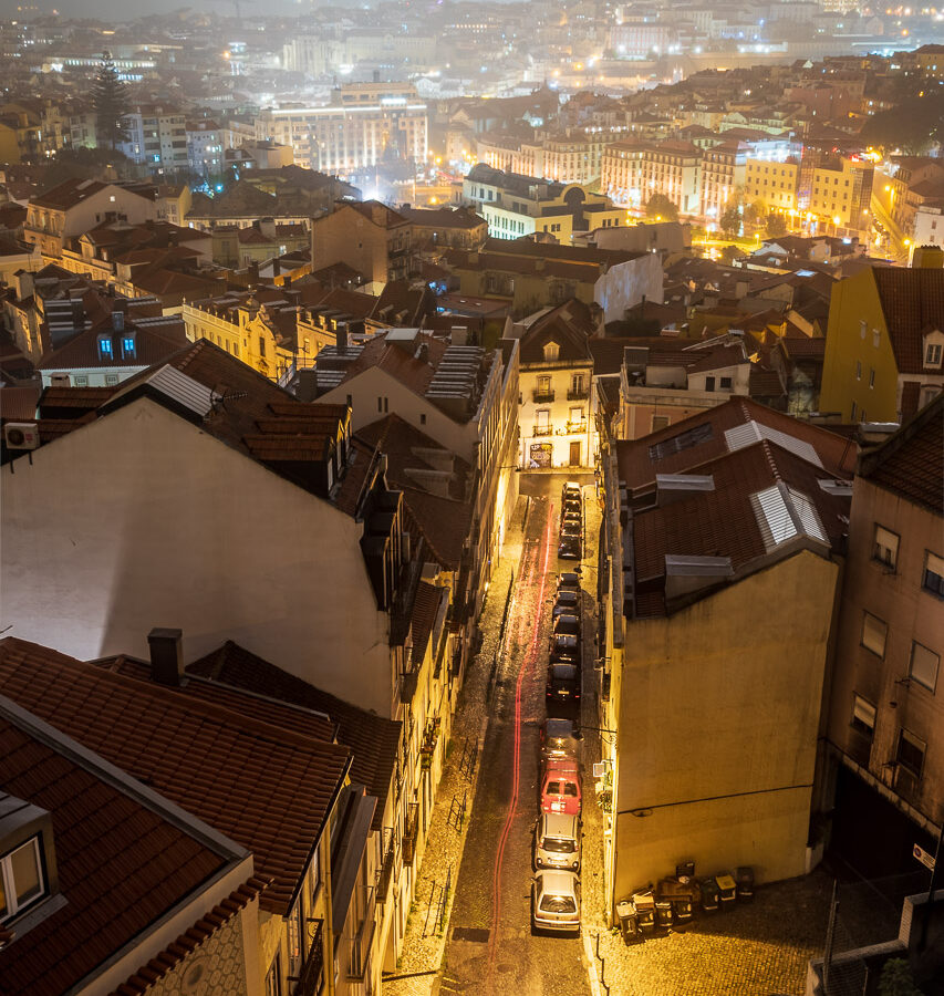A long exposure shot of Lisbon, with the trailing red brake lights of a car captured going down a nearby street.