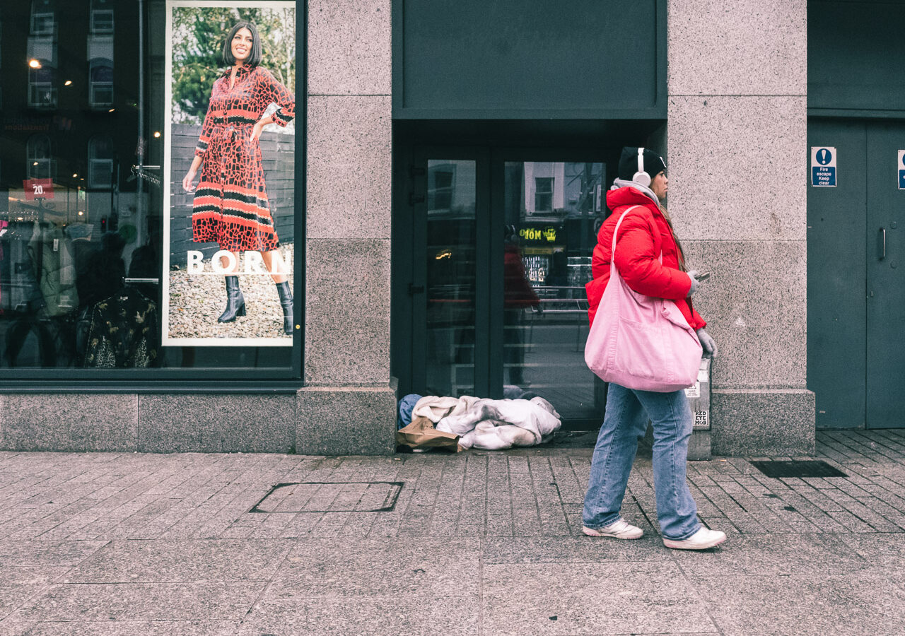 A woman walks past bed clothes in a doorway on a street while in a shop window a smiling woman looks on from a large poster.