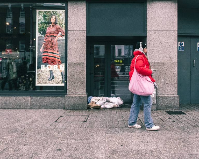 A woman walks past bed clothes in a doorway on a street while in a shop window a smiling woman looks on from a large poster.