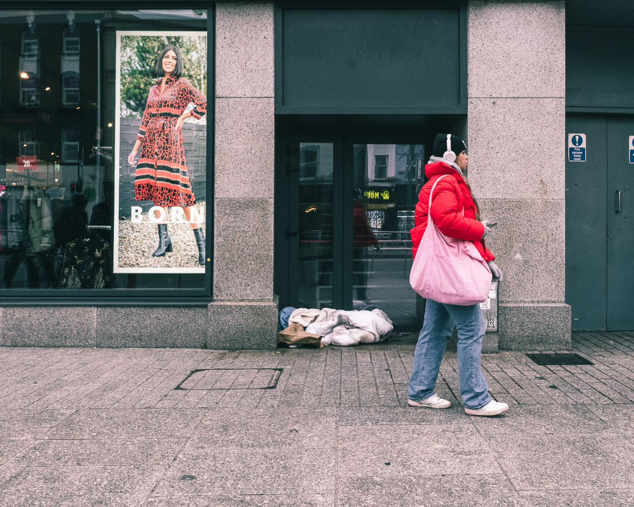 A woman walks past bed clothes in a doorway on a street while in a shop window a smiling woman looks on from a large poster.