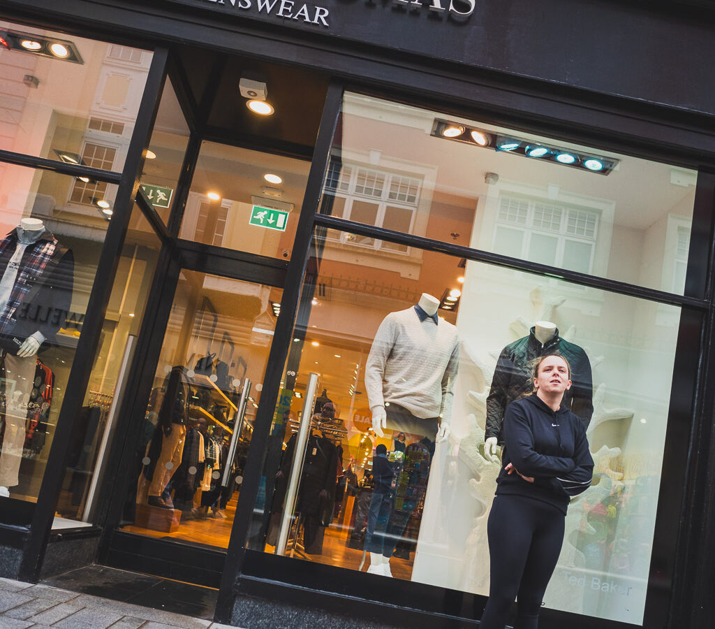 A woman stands in front of a Brown Thomas shop window, in front of a mannequin dressed in dark clothes, just like she is.