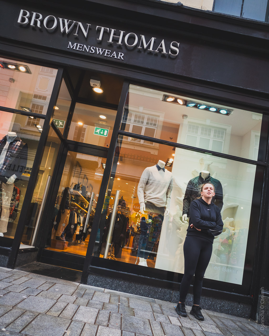 A woman stands in front of a Brown Thomas shop window, in front of a mannequin dressed in dark clothes, just like she is.